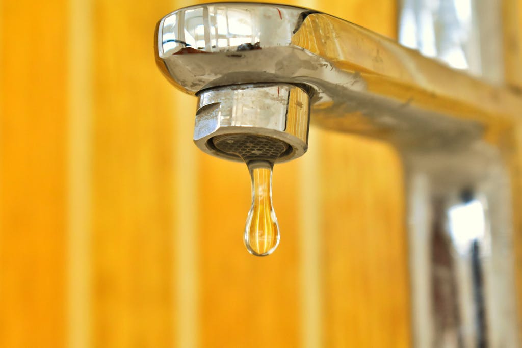 A close-up shot of a water droplet hanging from a stainless steel faucet, highlighting surface tension. Fixing a Leaky Faucet DIY Guide